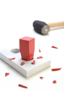 A square red peg hammered into a round hole surrounded by small pieces of the peg. Hammer in the background. On a white background